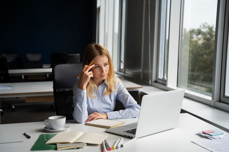 Mulher pensativa em frente ao computador, simbolizando o estresse causado pela cobrança excessiva no trabalho e a reflexão sobre como lidar com a situação.
