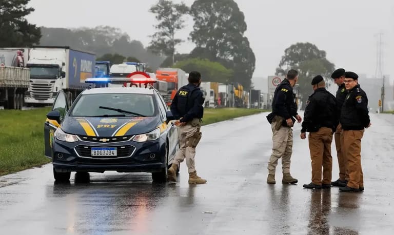 Policiais rodoviários federais durante operação em estrada chuvosa, com viatura e caminhões ao fundo.