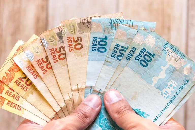 Bolsa Família Mãos segurando várias cédulas de real, incluindo notas de 20, 50 e 100 reais.