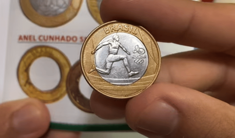 Moeda Olímpica de 1 Real: Atletismo A mão de uma pessoa segurando uma moeda olímpica do atletismo de 1 real, mostrando o salto com vara, com o catálogo de moedas ao fundo.