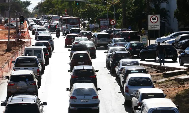Trânsito pesado em uma rua com várias faixas e carros estacionados ao longo do meio-fio, representando a circulação de veículos na cidade.