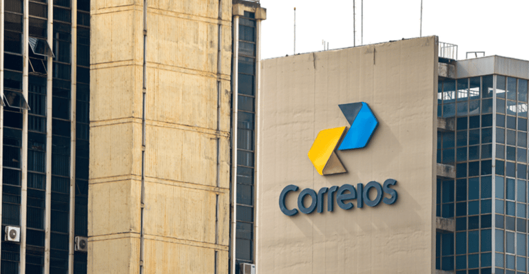 Fachada do edifício dos Correios em um dia de sol. O logotipo dos Correios, com três setas em amarelo, azul e cinza, está visível no prédio principal.
