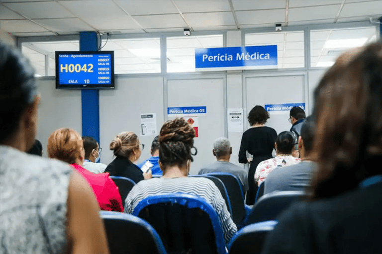 Pessoas sentadas aguardando atendimento em sala de perícia médica com painel eletrônico de senhas.