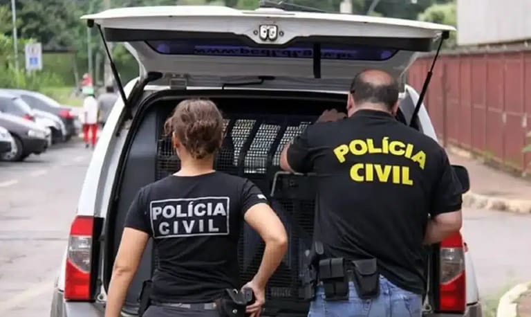 Dois policiais civis de costas com uniformes pretos próximos a viatura em operação policial
