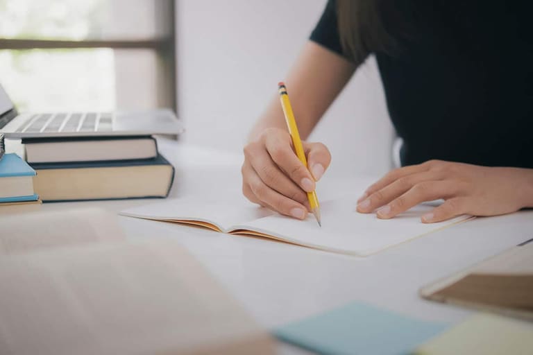 Pessoa estudando e escrevendo em um caderno com livros e notebook ao fundo.