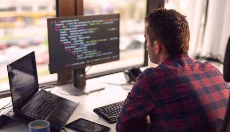 Jovem desenvolvedor de software de costas, trabalhando em frente a dois monitores exibindo código e utilizando um laptop em um escritório moderno, ilustrando o profissional e a carreira de tecnologia.