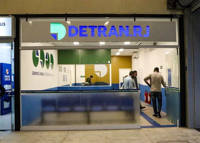 Fachada de um posto de atendimento do DETRAN RJ dentro de um shopping ou centro comercial. Pessoas aguardam ou conversam em balcões de atendimento, destacando-se a placa azul com o logo e o nome “DETRAN.RJ” em letras maiúsculas.