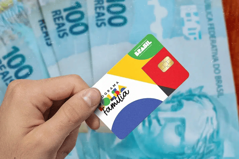 Mão segurando cartão do Programa Bolsa Família com cédulas de 100 reais ao fundo