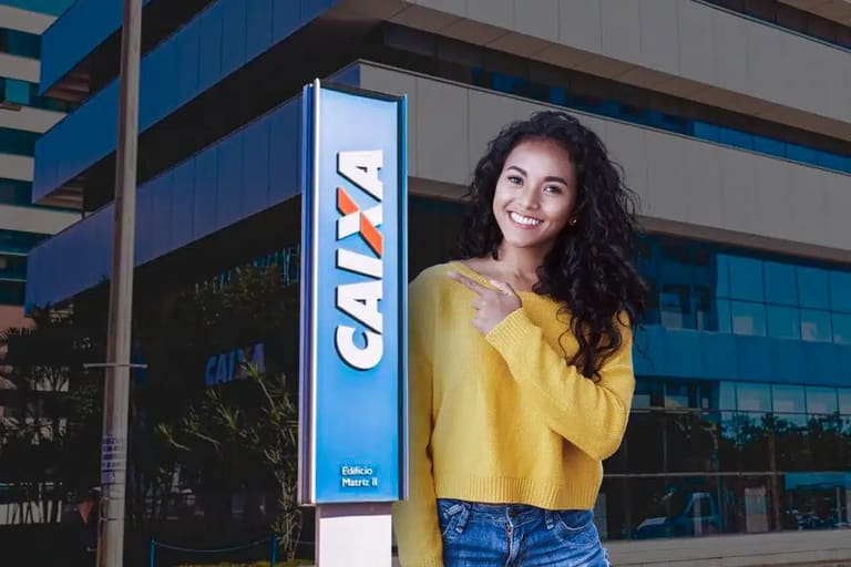 Mulher jovem e sorridente de suéter amarelo aponta placa azul da Caixa Econômica em frente a prédio corporativo.