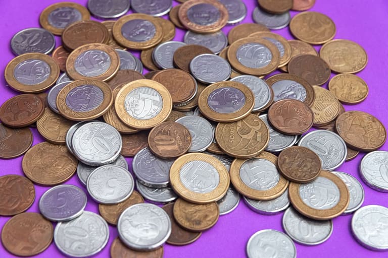 Descubra Moedas Raras: Do Real ao Tesouro de R$ 6.000 Coleção de moedas brasileiras de diferentes valores e anos, espalhadas sobre um fundo roxo, ilustrando a busca por moedas raras e valiosas.