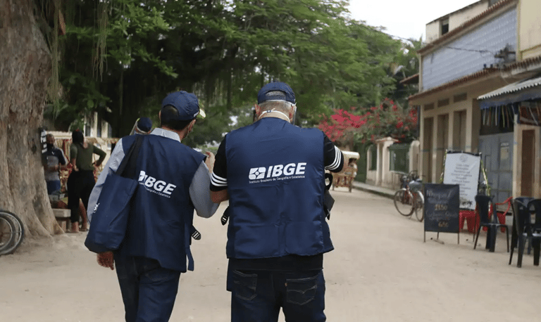 Dois agentes de pesquisa do IBGE usando coletes azuis identificados caminhando por rua durante trabalho de campo.