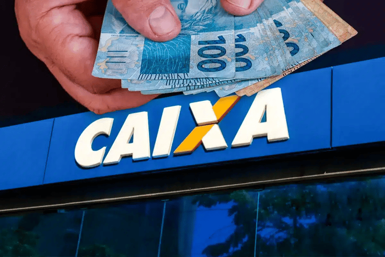 Mão segurando cédulas de real em frente a agência da Caixa Econômica Federal anunciando pagamento de R$ 1.110 neste fim de ano.