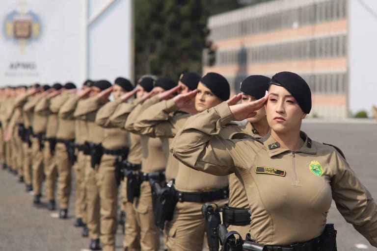 Policiais militares femininas alinhadas em formação realizando saudação com uniforme bege e boina preta