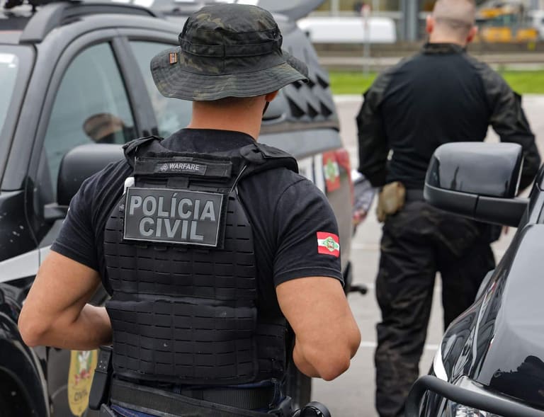 Agentes da Polícia Civil com colete tático preto diante de viaturas durante ação policial.