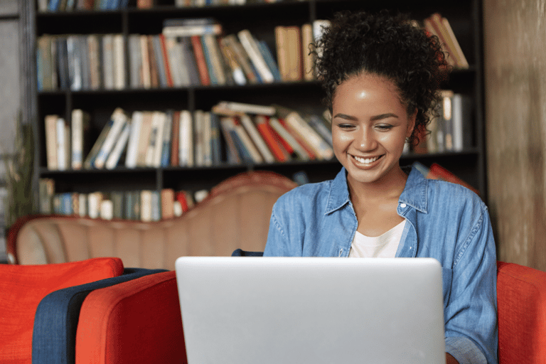 Jovem sorridente usando notebook sentada em poltrona vermelha com estante de livros ao fundo