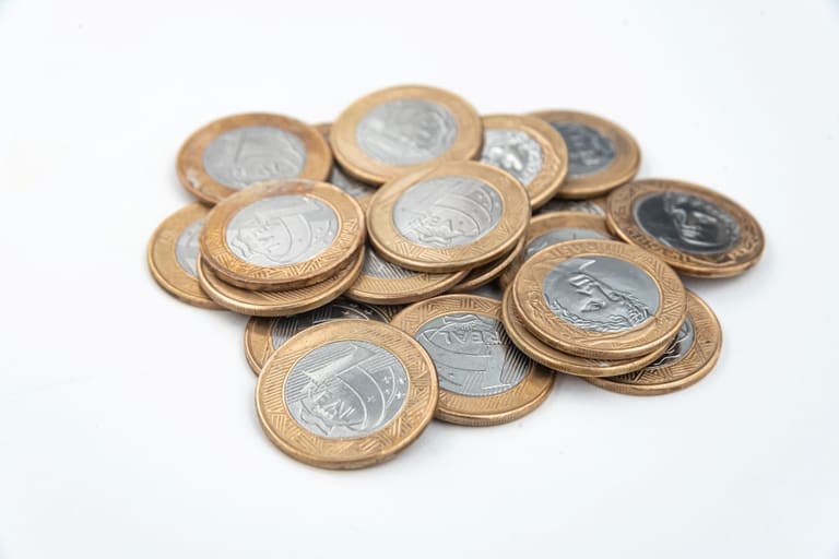 Pilha de moedas de 1 real de faces variadas, incluindo a efígie da República e a Cruz do Sul, sobre uma superfície branca limpa.