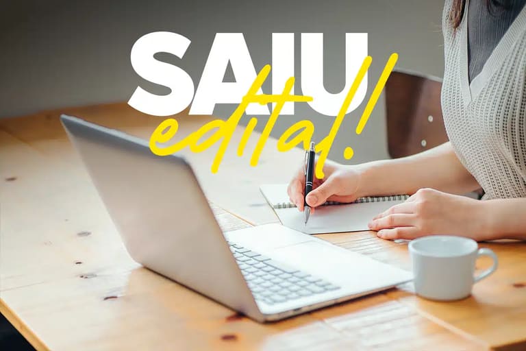 Saiu edital: concurso prefeitura Pessoa escrevendo em caderno ao lado de notebook em mesa de madeira, com texto SAIU Edital em branco e amarelo.