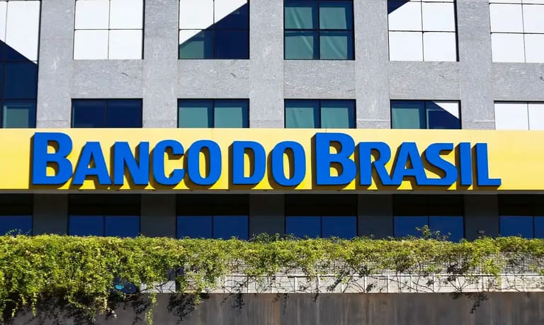 Placa amarela com letras azuis do Banco do Brasil em fachada de prédio moderno cinza com janelas espelhadas