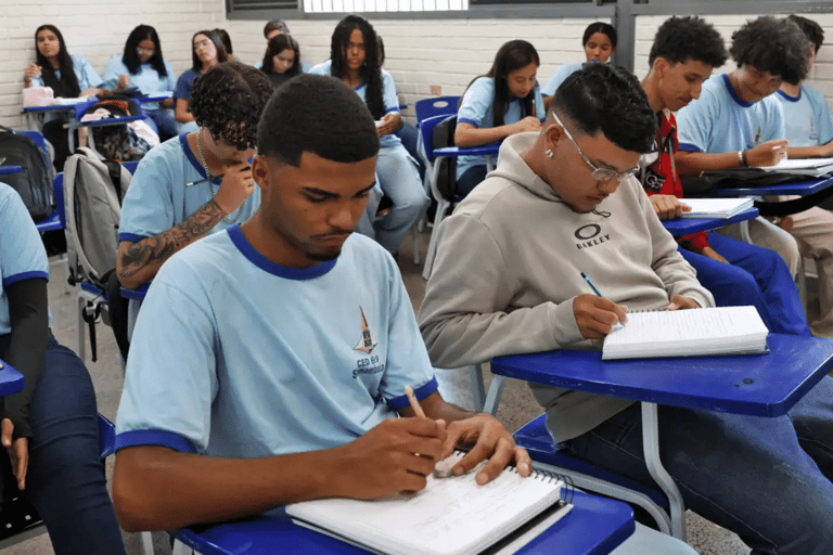 Jovens estudantes sentados em carteiras azuis escrevendo em cadernos em uma sala de aula com paredes de tijolos brancos