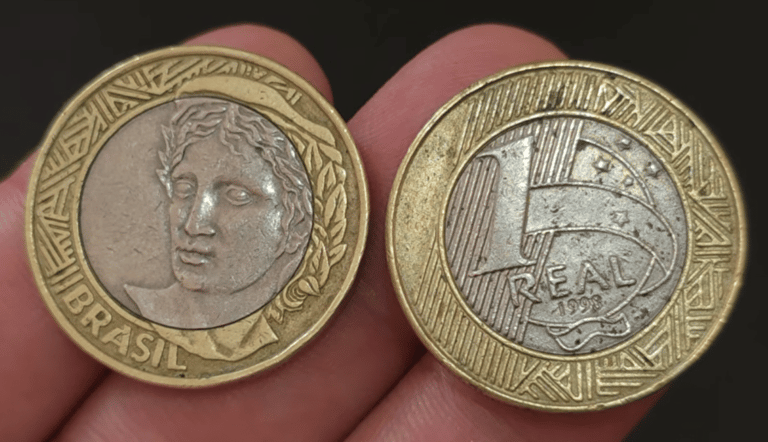 Duas moedas de 1 real de 1998 lado a lado; a da esquerda mostra o anverso com a efígie da República e a da direita exibe o valor de face, o ano 1998 e o núcleo prateado.