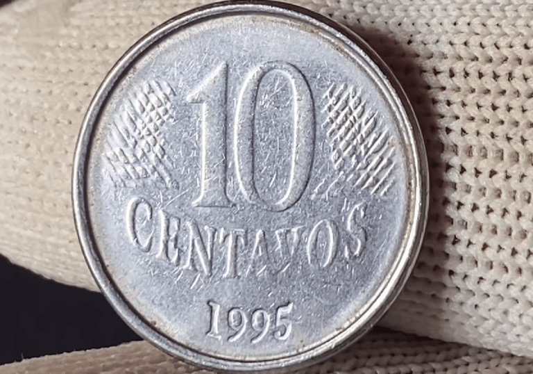 Close detalhado de diversas moedas brasileiras de um real e de centavos empilhadas sobre uma mesa de madeira, com foco em uma moeda de um real em destaque.