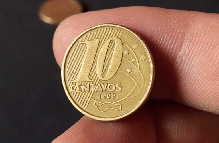 Close-up de dedos segurando uma moeda de 10 centavos de Real, cor dourada, com o ano de 1999 visível na parte inferior. O fundo é escuro e desfocado.