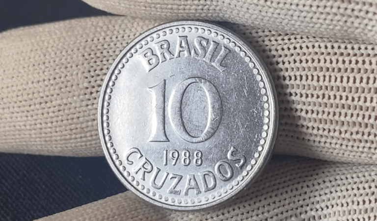 Close-up de uma moeda prateada de 10 Cruzados do ano de 1988, sendo segurada por uma mão com luva de tecido branco. A moeda exibe a inscrição "BRASIL", o valor "10" e "CRUZADOS" em destaque.
