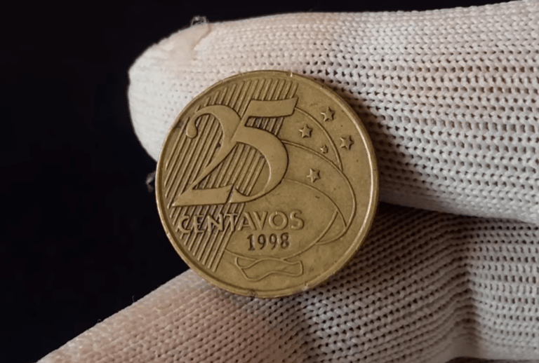 Close-up de uma moeda de 25 centavos de 1998 sendo segurada por dedos com luvas brancas, focando no reverso onde a data aparece marcada sob os ramos.