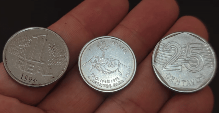 Três moedas brasileiras raras estendidas sobre a palma de uma mão: uma de 1 Real de 1994, uma comemorativa da FAO com o desenho de mãos segurando uma planta e uma de 25 centavos de 1995.