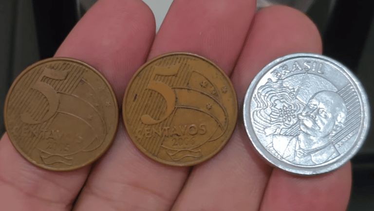 Três moedas de cinco centavos raras sendo seguradas entre os dedos: duas de cor bronze (anos 2005 e 2006) e uma prateada (híbrida de 2012).