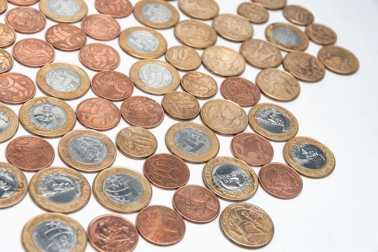 Vista superior de várias moedas brasileiras do Plano Real, incluindo moedas de 1 Real, 50, 25, 10 e 5 centavos, espalhadas de forma aleatória sobre uma superfície branca.