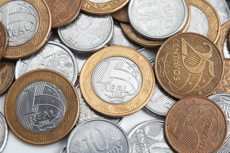 Uma pilha mista de moedas do Real brasileiro de diversos valores, incluindo moedas de 1 Real, 25 centavos, 10 centavos e 5 centavos, espalhadas sobre uma superfície clara.