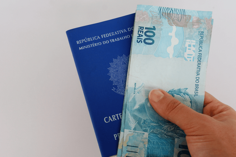 Mão segurando uma nota de 100 reais em frente a uma carteira de trabalho azul do Brasil