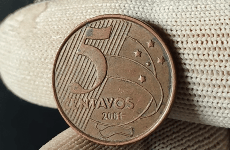 Close de uma moeda de 5 centavos do ano 2001 sendo segurada por dedos que usam luvas de tecido branco. A moeda de cobre mostra o número 5, a palavra "CENTAVOS" e o ano 2001 em destaque.