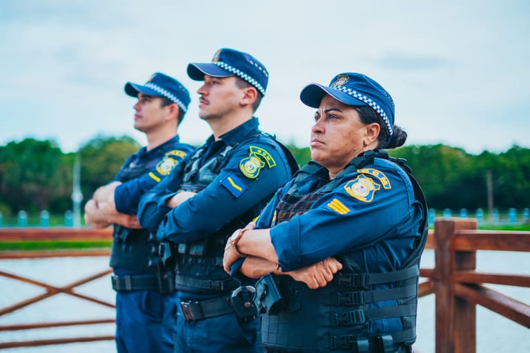 Três guardas municipais uniformizados de braços cruzados em área aberta próxima à água