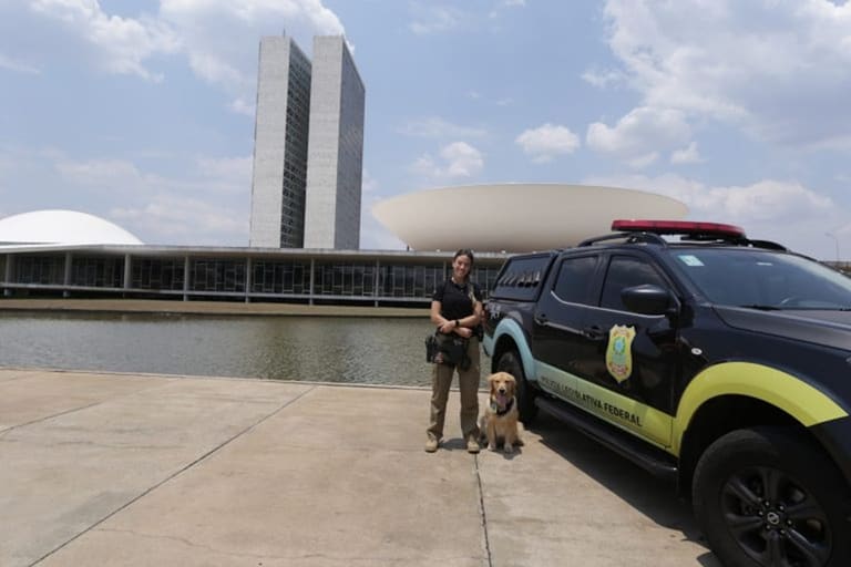 Policial legislativa federal com cão farejador ao lado de viatura em frente ao Congresso Nacional em Brasília