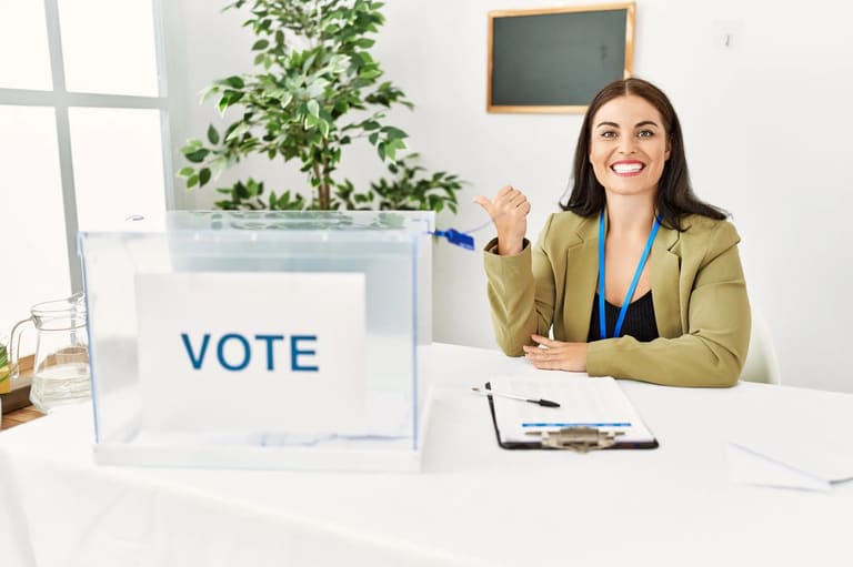 Mesária sorrindo e apontando com o polegar ao lado de uma urna eleitoral nas Eleições de 2026.