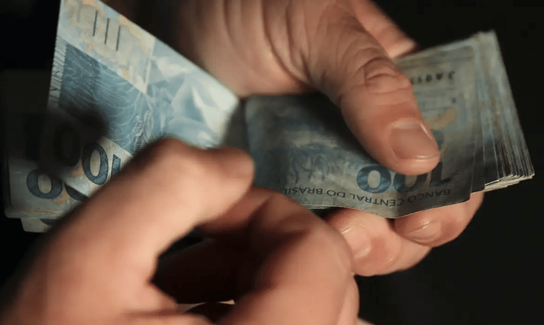 Mãos contando notas de cem reais do Banco Central do Brasil representando o microcrédito para inscritos no CadÚnico