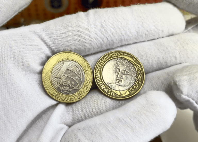 Duas mãos usando luvas brancas de tecido seguram duas moedas de 1 real de 1998. A moeda da esquerda mostra o valor "1 REAL" e o ano "1998" no centro de cupro-níquel, cercado por um anel de alpaca dourado. A moeda da direita mostra a efígie da República voltada para a esquerda.