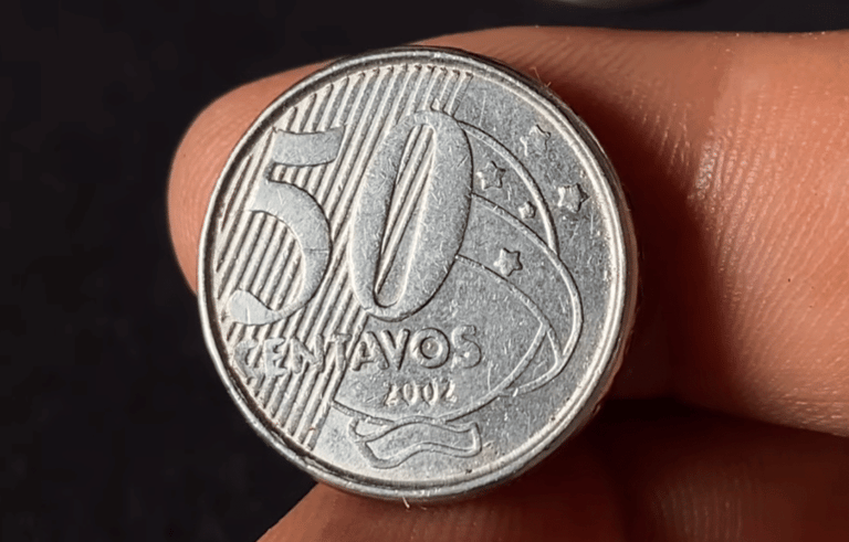 Close-up detalhado de uma moeda de 50 centavos de real do ano 2002, fabricada em aço inoxidável, sendo segurada entre os dedos contra um fundo escuro.
