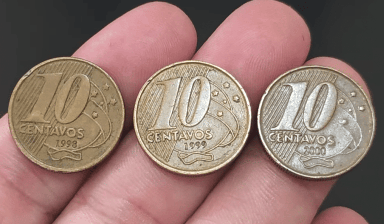 Close de uma mão segurando três moedas de 10 centavos da segunda família do Real, alinhadas lado a lado. As moedas exibem claramente os anos de cunhagem 1998, 1999 e 2000, da esquerda para a direita.