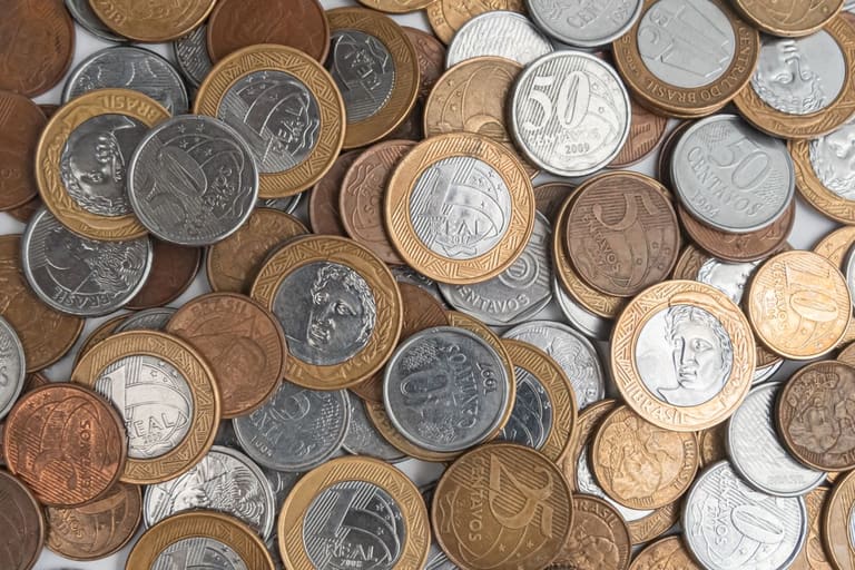 Diversas moedas do Plano Real espalhadas e sobrepostas, incluindo moedas de 1 Real, 50 centavos e 10 centavos de diferentes anos e metais.