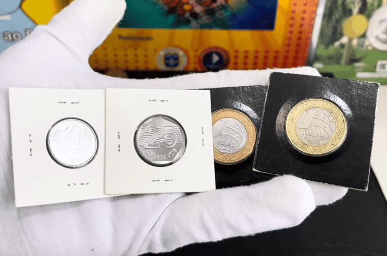 Mão usando luva branca de colecionador segurando quatro moedas raras do Real protegidas por suportes de papelão (coin holders). Estão visíveis as moedas de 10 centavos, 25 centavos e duas de 1 Real.
