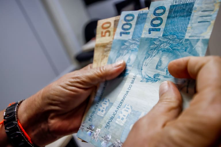 13° salário do INSS Mãos contando notas de R$ 100 e R$ 50 em ambiente interno