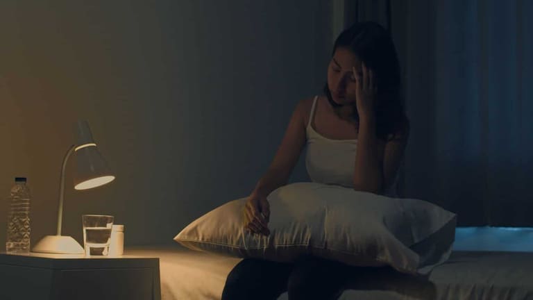 Mulher jovem asiática com dor de cabeça à noite, sofrendo de insônia enquanto tenta dormir em um ambiente com pouca luz. Estudo científico revela que dormir tarde pode afetar o coração a longo prazo.