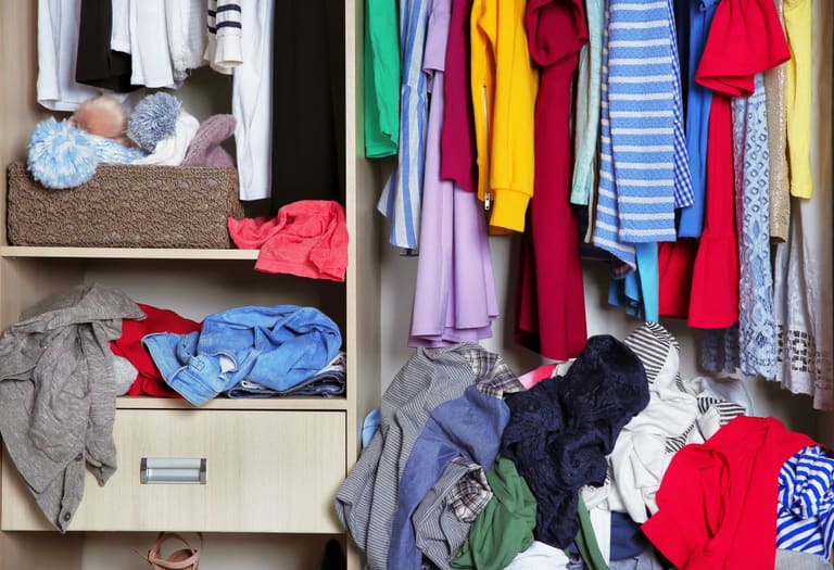 Interior de guarda-roupa com roupas penduradas e várias peças acumuladas e desorganizadas nas prateleiras e no chão.