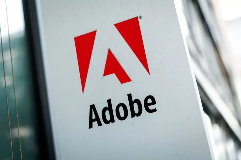 Logotipo da Adobe com o ícone "A" estilizado em vermelho e o nome da marca em preto, exibido em uma placa branca na fachada de um edifício moderno.