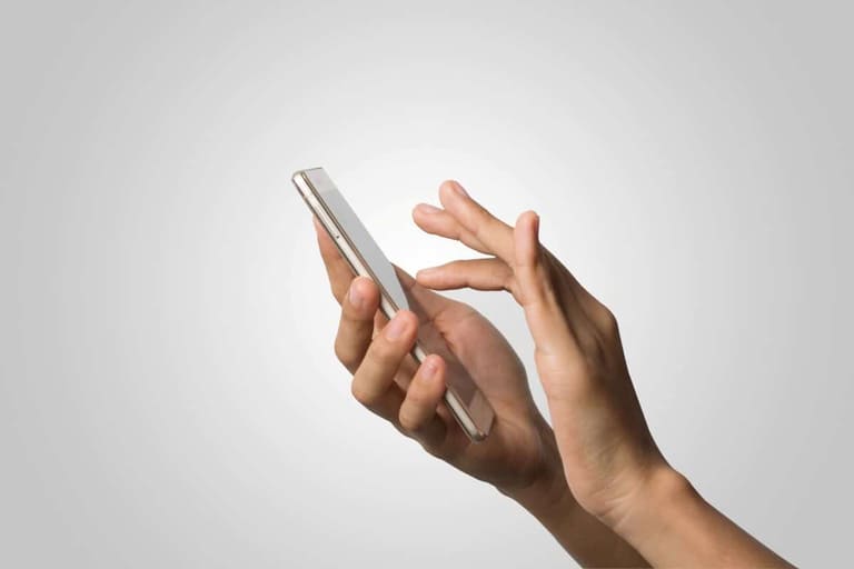 Close-up de mãos segurando um smartphone fino de cor dourada clara, com o dedo indicador tocando a tela, contra um fundo cinza neutro e minimalista.