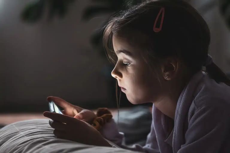 "Menina pequena usando um smartphone na cama, focada na tela, representando a questão do uso de dispositivos por crianças.