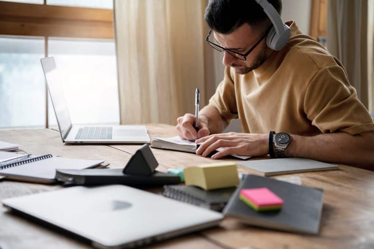 Homem estudando em casa com notebook e caderno enquanto se prepara para decisões de carreira entre faculdade e concursos públicos.